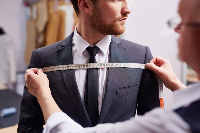 A tailor measuring the chest width of a young bearded man, who is wearing a custom made jacket, shirt and tie.
