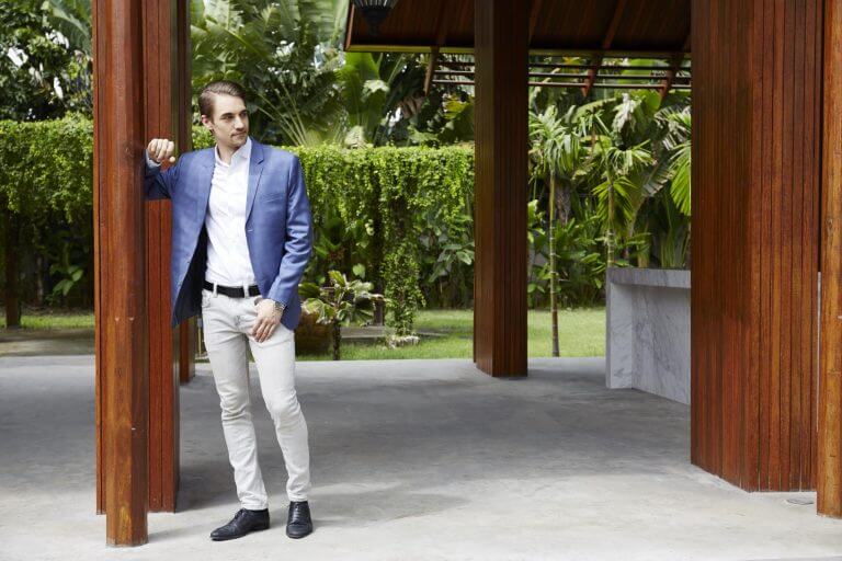 A man standing outside a house in front of garden with palm trees. He is wearing white tailored trousers, a white shirt and a tailored blue jacket.