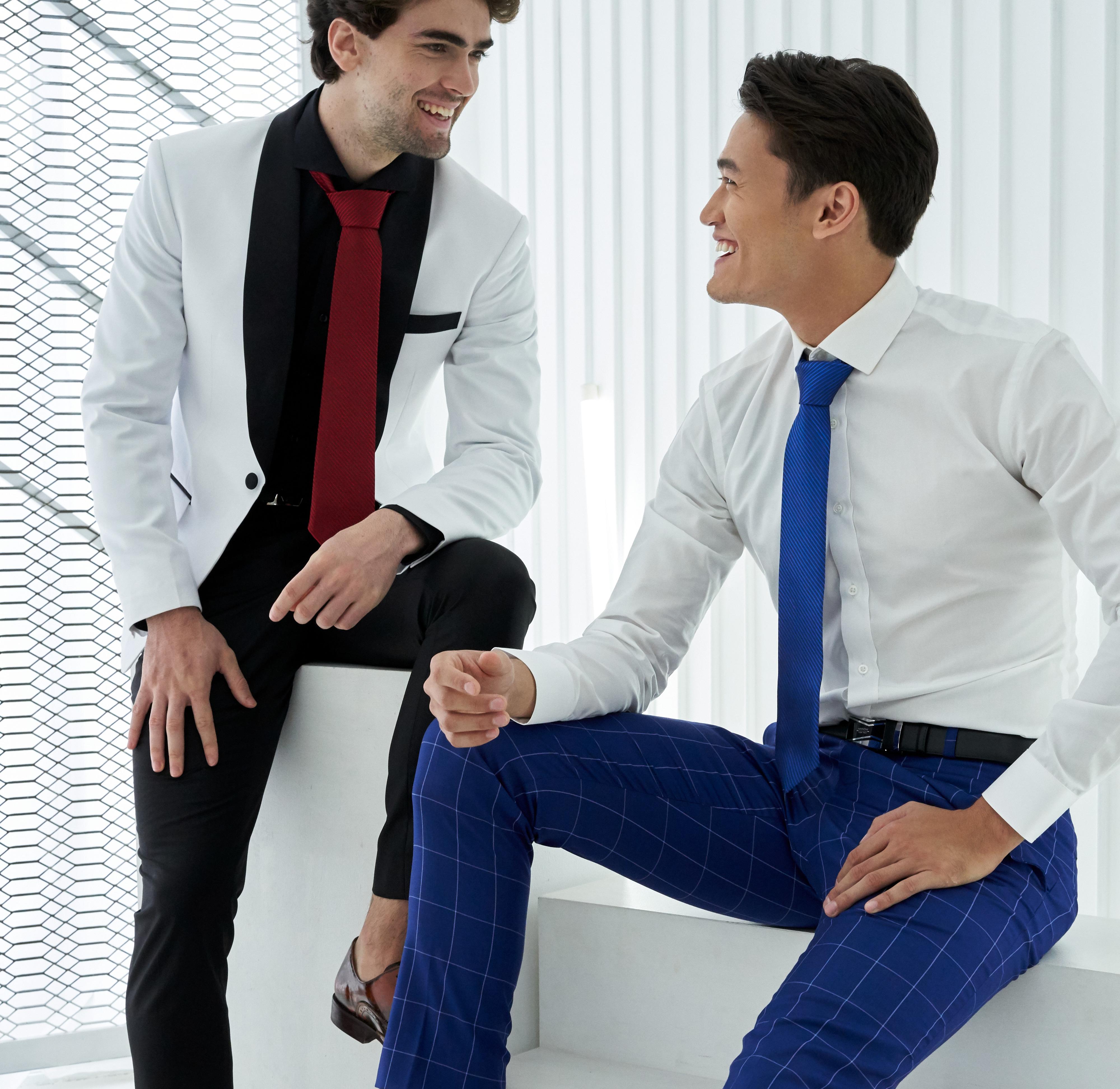 Two white men talking to each other. One is wearing a black tailored shirt with a custom made white jacket and a red tie. The other is wearing blue checkered pants, a white shirt and a blue tie.